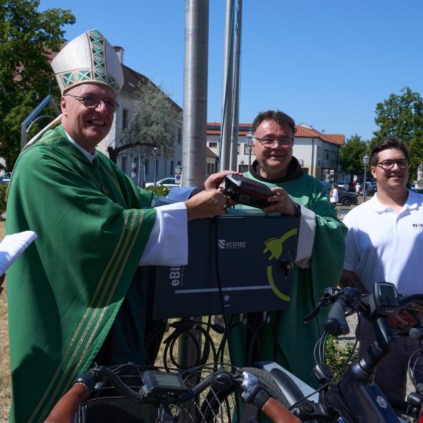 Ein Bischof segnet ein Fahrrad-Ladegerät, umgeben von Helfern und Fahrrädern im Freien.