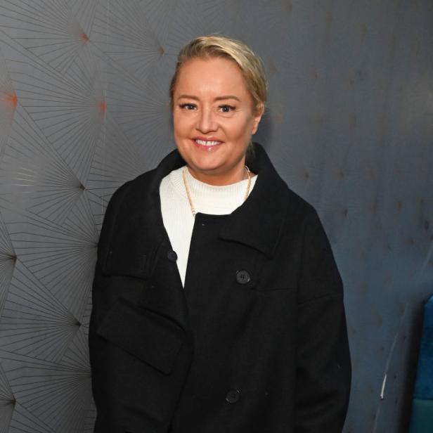 Lucy Davis lächelt in schwarzem Mantel vor einer grauen Wand.