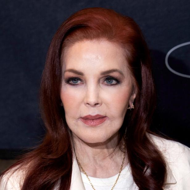 Priscilla Presley mit rötlichem Haar und weißem Blazer vor dunklem Hintergrund.