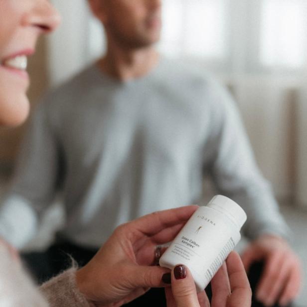 Frau hält BIOGENA Osteo Calbon Komplex Gold Dose in der Hand, Mann sitzt im Hintergrund im Schneidersitz.