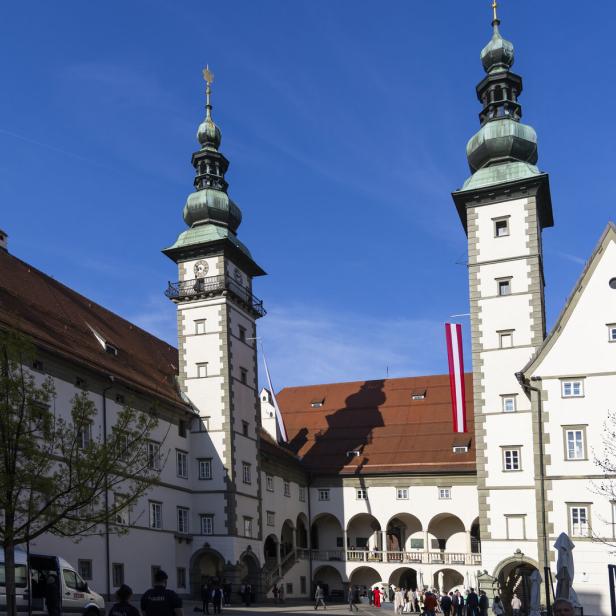 Historischer Innenhof mit zwei Türmen, Arkaden und Menschen unter blauem Himmel.