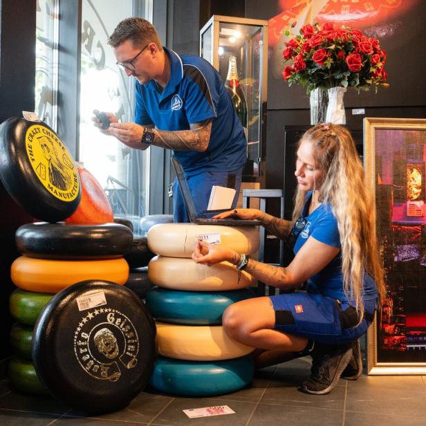 Zwei Personen in blauer Arbeitskleidung inventarisieren in einem Geschäft mehrere farbige Käselaibe von The Crazy Cheese Manufacture.