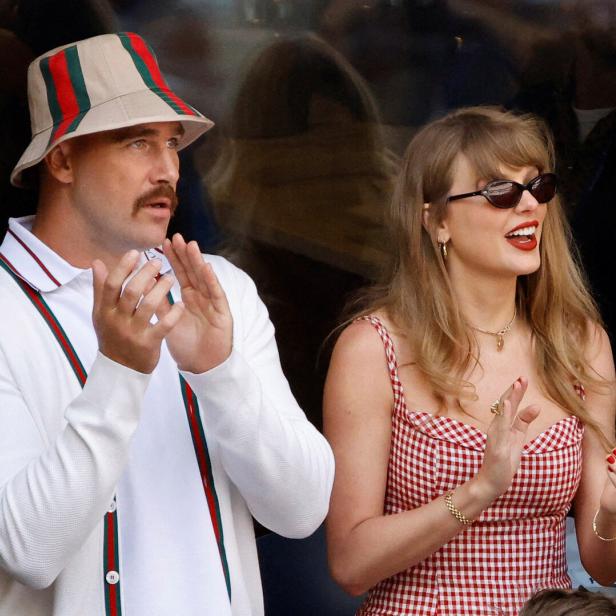 Travis Kelce mit Fischerhut und Taylor Swift in einem rot-weiß karierten Kleid klatschen gemeinsam auf einer Tribüne.
