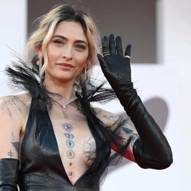 Paris Jackson winkt in einem schwarzen Lederkleid mit Federkragen und sichtbaren Tattoos auf dem roten Teppich.