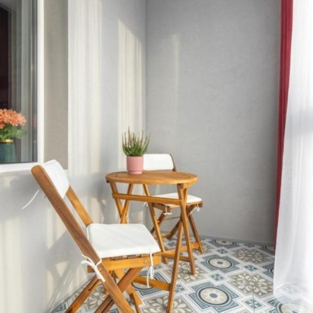 Kleiner Balkon mit zwei Holzstühlen mit weißen Sitzkissen und einem runden Holztisch auf gemustertem Boden.