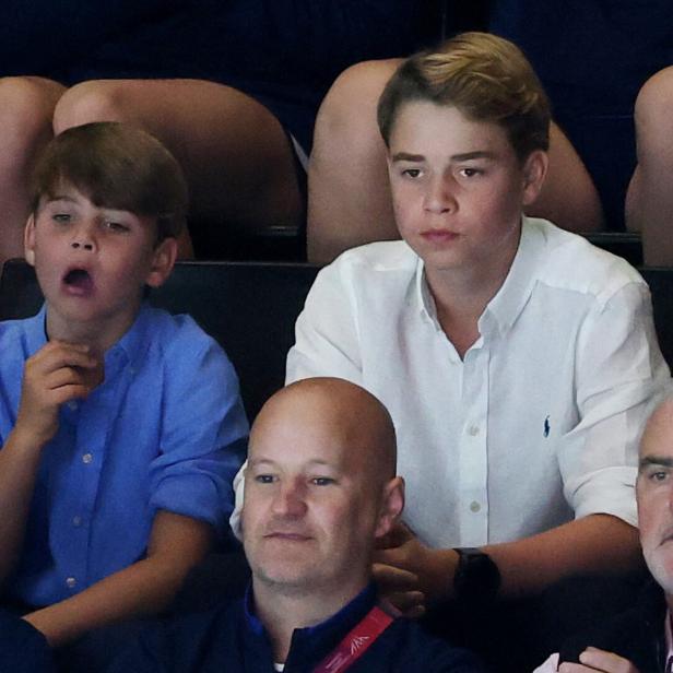 Prinz George und Prinz Louis sitzen bei einer Veranstaltung auf der Tribüne, wobei Louis gähnt und George aufmerksam nach vorne blickt.