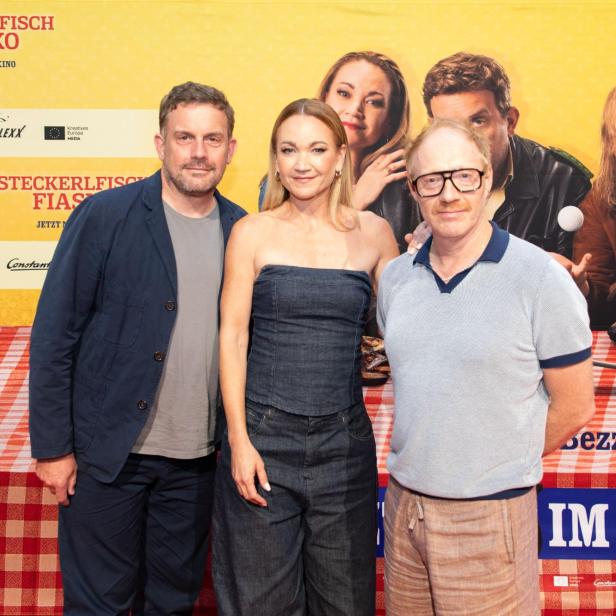 Sebastian Bezzel, Lisa Maria Potthoff und Simon Schwarz posieren vor dem „Steckerlfischfiasko“-Backdrop bei der Premiere in Wien.