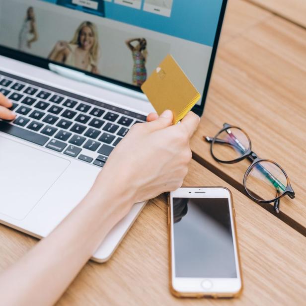Person hält goldene Kreditkarte über Laptop-Tastatur auf hellem Holztisch mit Smartphone und Brille daneben.