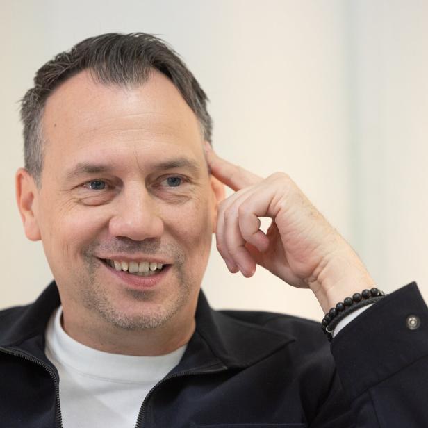 Ein lächelnder Sebastian Fitzek mit kurzem Haar trägt eine schwarze Jacke über einem weißen T-Shirt und hält seine Hand nachdenklich an die Schläfe.