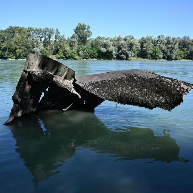 Ein verrostetes Wrackteil ragt aus dem Wasser eines Flusses vor einem bewaldeten Ufer hervor.