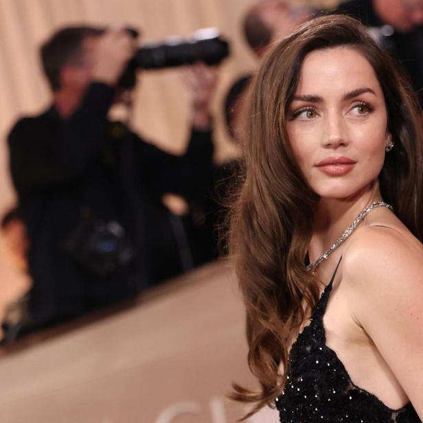 Ana de Armas blickt mit langem, braunem Haar und in einem schwarzen Abendkleid vor einer Kulisse mit Fotografen zur Seite.