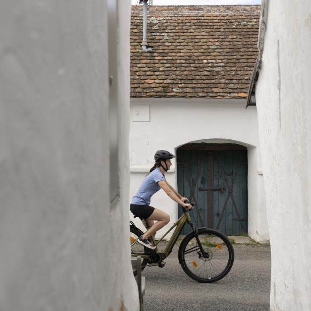 Fahrradfahrerin mit Helm fährt durch eine enge Gasse vor einem weißen Haus mit blauem Tor.