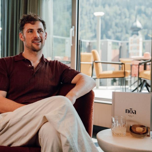 Ein Mann in einem bordeauxroten Poloshirt sitzt entspannt in einem Sessel neben einer Menükarte mit der Aufschrift „nōa“ in einem hellen Raum.