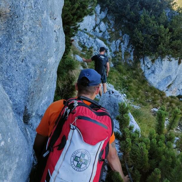 Zwei Wanderer klettern auf einem schmalen Bergpfad zwischen Felsen und Latschen.