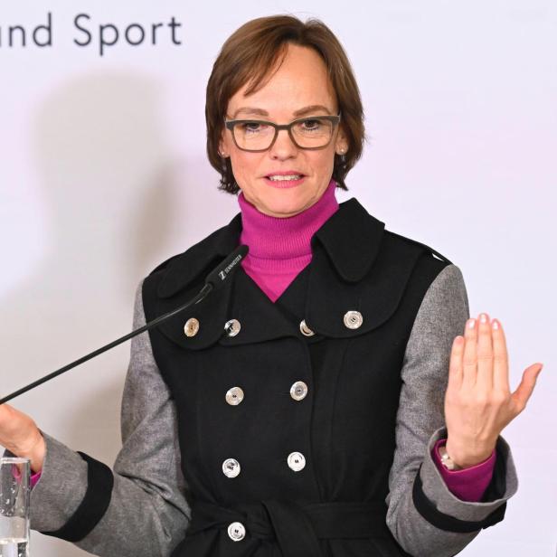 Frau mit Brille spricht am Mikrofon und hebt beide Hände vor einem hellen Hintergrund.
