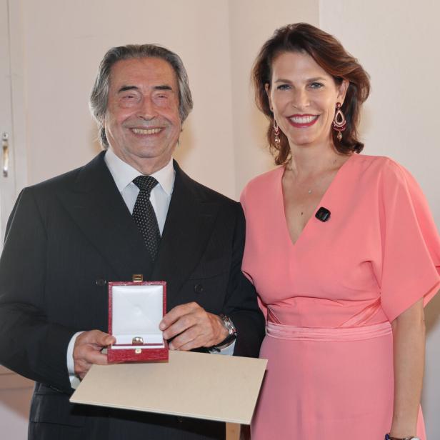 Riccardo Muti hält ein Schmuckkästchen mit einem Ring in der Hand und lächelt neben einer Frau in einem lachsfarbenen Kleid.