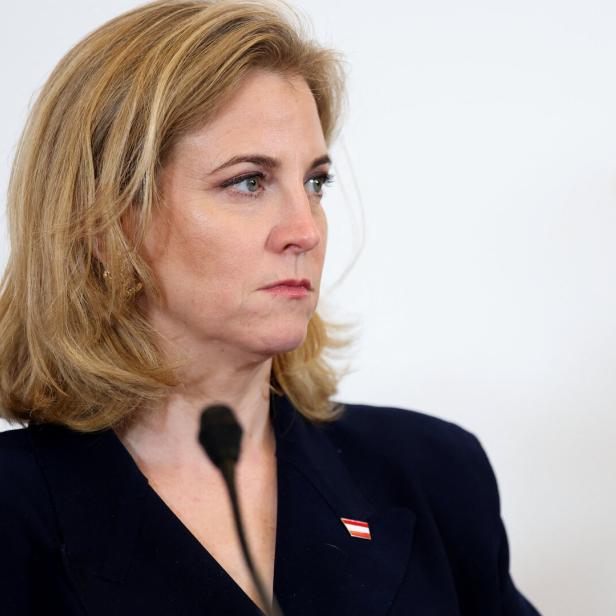 Eine blonde Frau im dunklen Blazer spricht an einem Mikrofon vor hellem Hintergrund.