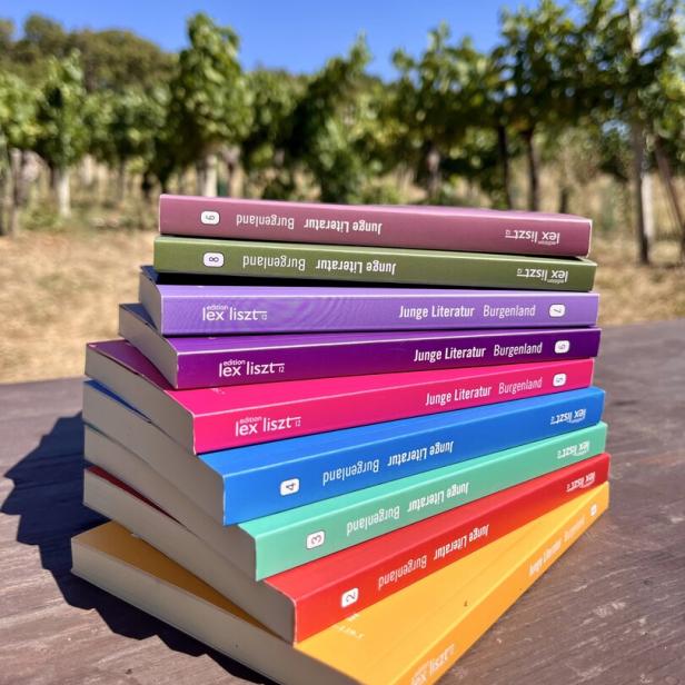 Ein Stapel bunter Bücher der Reihe „Junge Literatur Burgenland“ der edition lex liszt 12 liegt auf einem Holztisch vor einem Weinberg.