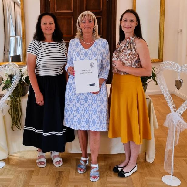 Drei Frauen (in der Mitte Stadtchefin Böhm) stehen nebeneinander, wobei die mittlere eine Urkunde der Stadt Neusiedl am See hält, flankiert von zwei weißen Herz-Dekorationen.