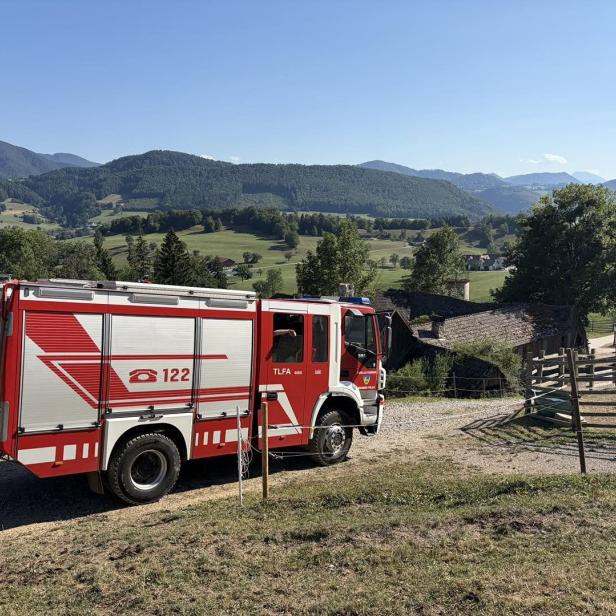 Ein rotes Feuerwehrfahrzeug steht auf einem Feldweg vor einer ländlichen Bergkulisse.