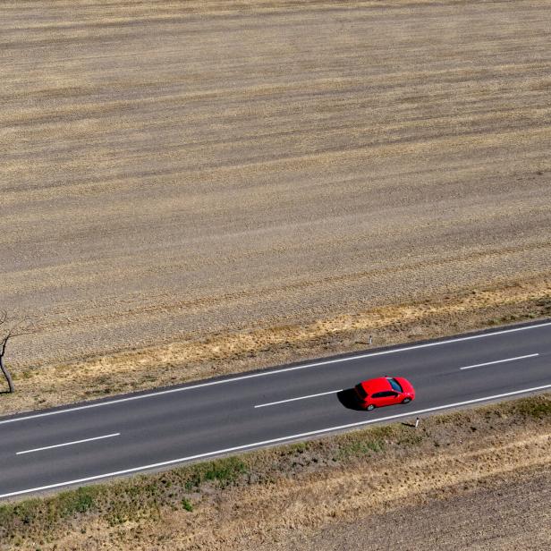 Ein rotes Auto fährt auf einer einsamen Landstraße durch eine weitläufige, trockene Ackerlandschaft.