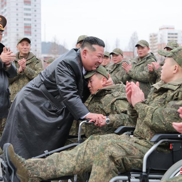 Kim Jong-un umarmt einen sitzenden Soldaten, umgeben von applaudierenden Militärangehörigen im Freien.