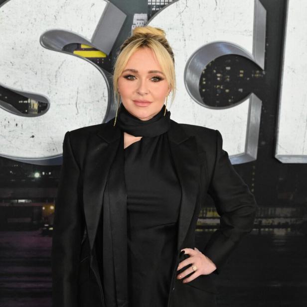 Hayden Panettiere in schwarzem Outfit mit Mantel und hochgesteckten Haaren vor einer Filmkulisse.