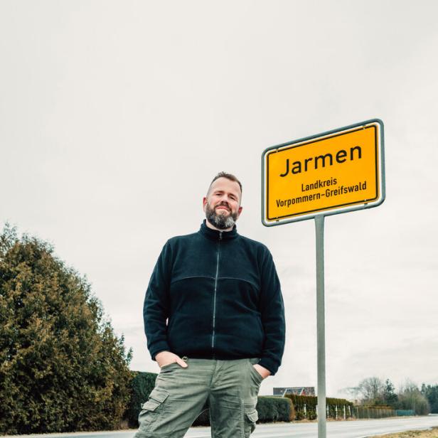 Ein bärtiger Mann steht neben einem gelben Ortseingangsschild der Stadt Jarmen im Landkreis Vorpommern-Greifswald an einer Landstraße.