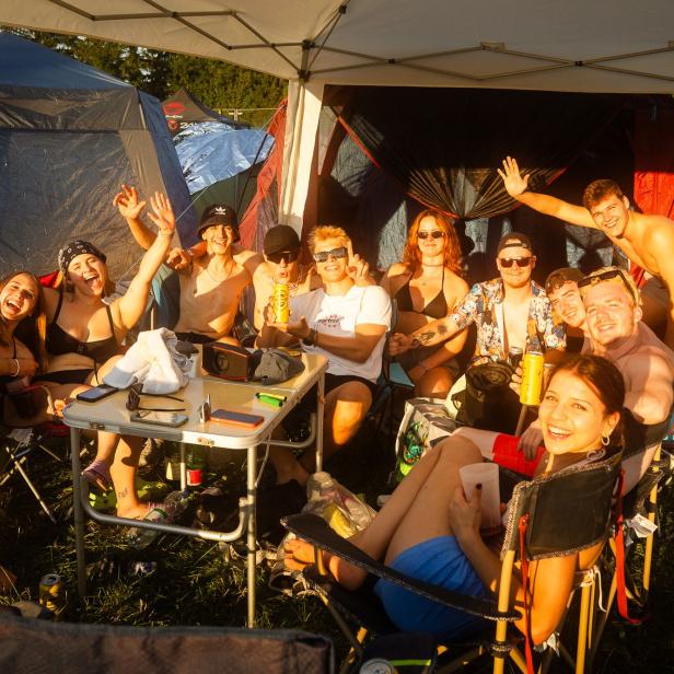 Festivalbesucherinnen und -besucher sitzen lachend im Campingbereich von Frequency zwischen Zelten und Campingstühlen.