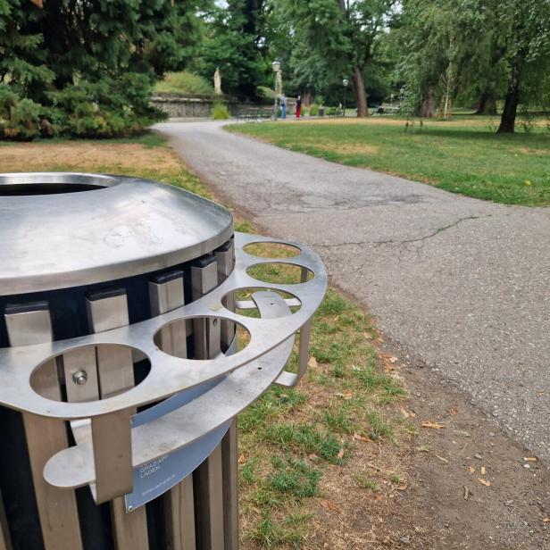 Ein Parkweg führt an einem großen Metall-Mülleimer vorbei, im Hintergrund stehen Bäume und vereinzelte Spaziergänger.