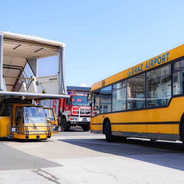 Gelbes Flughafen-Vorfeldfahrzeug und Bus am Graz Airport, dahinter ein Feuerwehrauto.