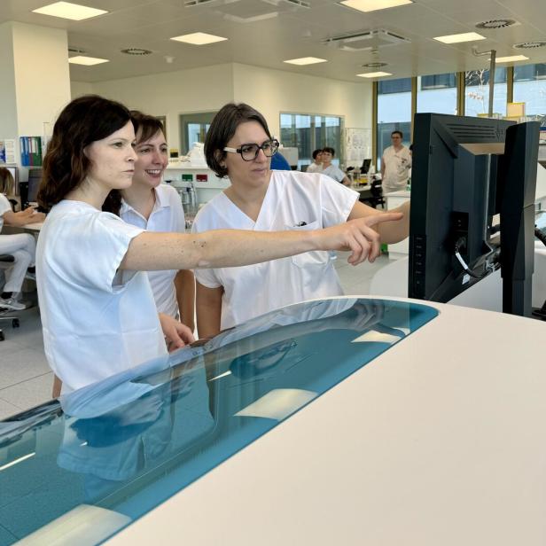 Drei Personen in weißen Kitteln schauen und zeigen in einem Labor auf einen Monitor.