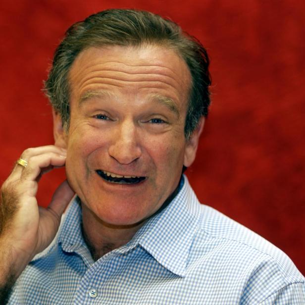 Robin Williams lächelt vor rotem Hintergrund und berührt sich am Ohr.