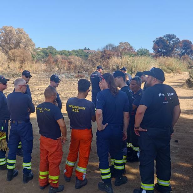 Mehrere Feuerwehrleute stehen in einer kargen Landschaft im Kreis.