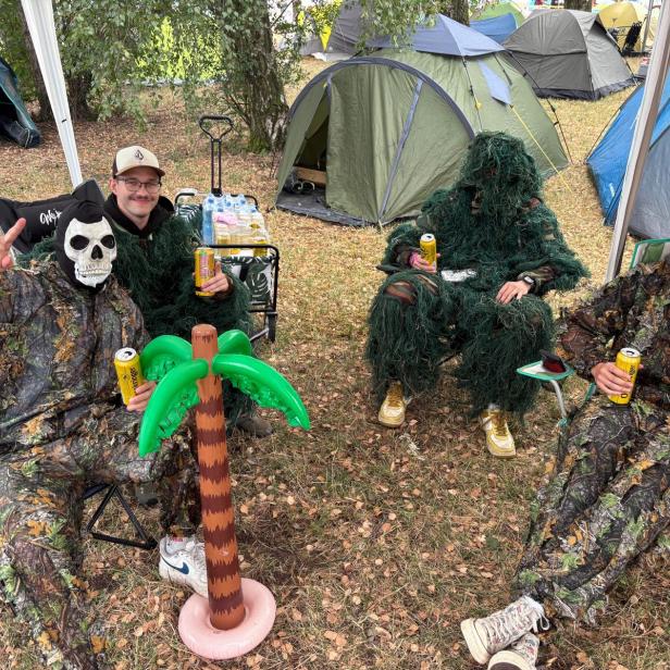 Vier als Waldmenschen verkleidete Frequency-Besucher sitzen mit Bierdosen vor Zelten, neben einer Plastikpalme auf dem Campingplatz.