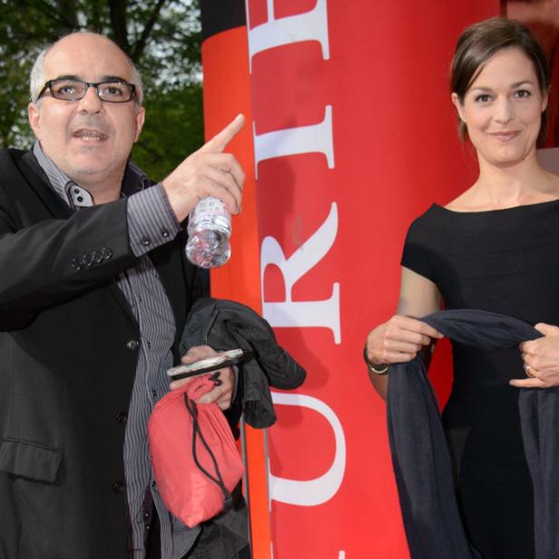 Ein Mann mit Brille und eine Frau in einem schwarzen Kleid posieren vor einer roten Wand.