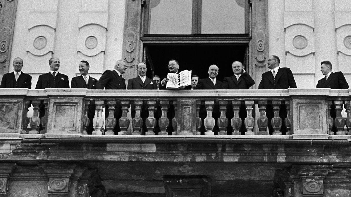 Eine Gruppe von Männern steht auf einem Balkon und blickt auf eine Menschenmenge.