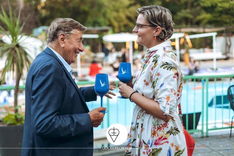 Ein Mann und eine Frau stehen mit blauen Gläsern am Pool und unterhalten sich.