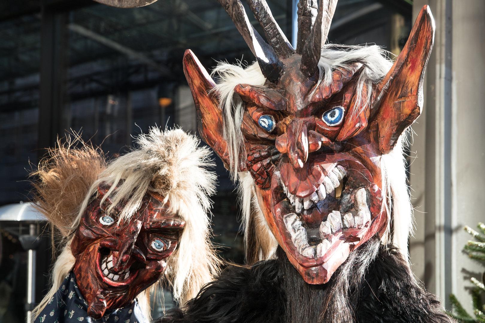 Zwei Krampusmasken mit Hörnern, Fell und gruseligen Gesichtern.