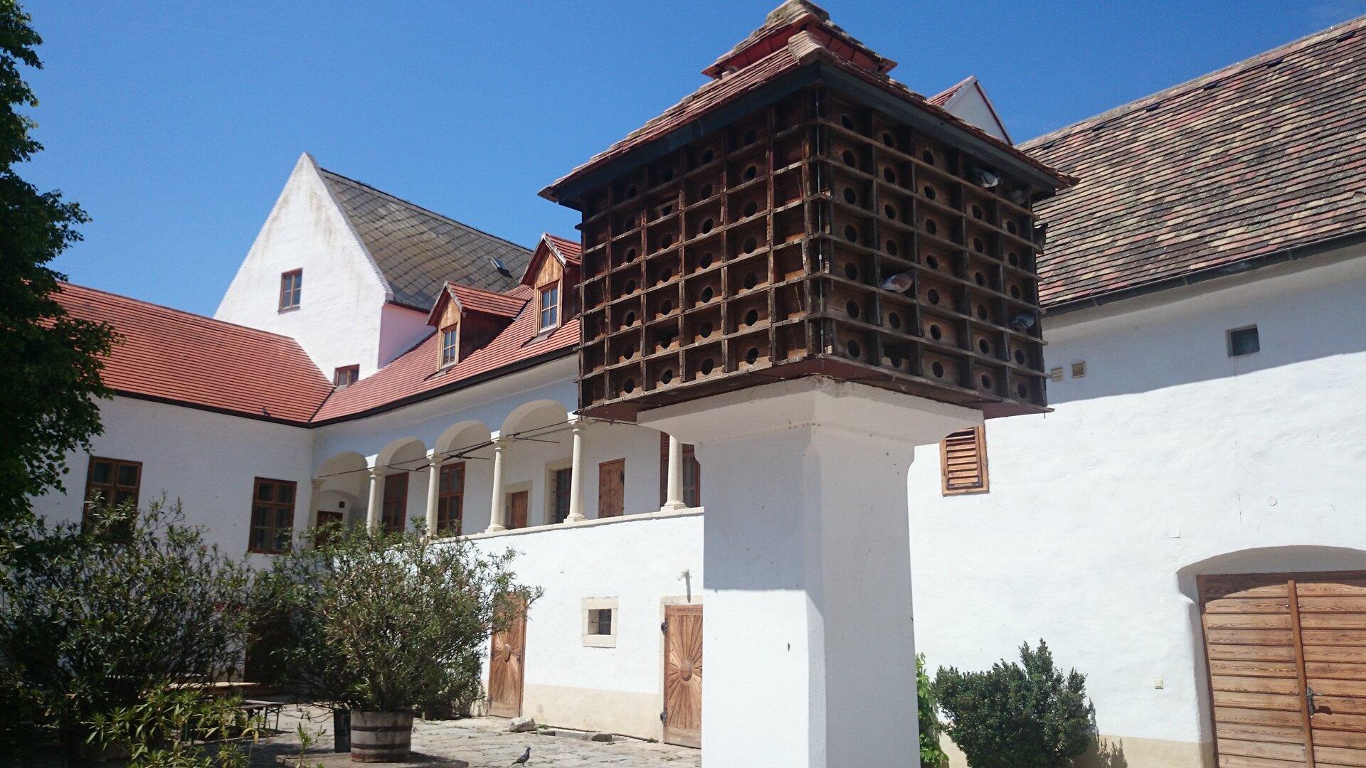 Ein großer hölzerner Taubenschlag steht auf einem weißen Sockel in einem sonnigen Innenhof mit historischen Gebäuden.