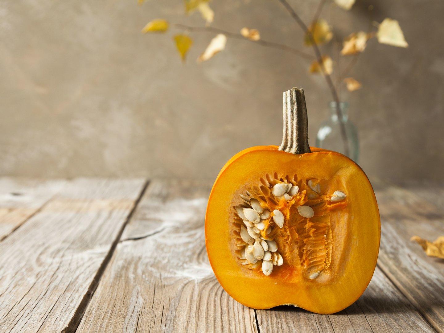 Cut ripe orange pumpkin with seeds and fallen leaves on pastel brown background.