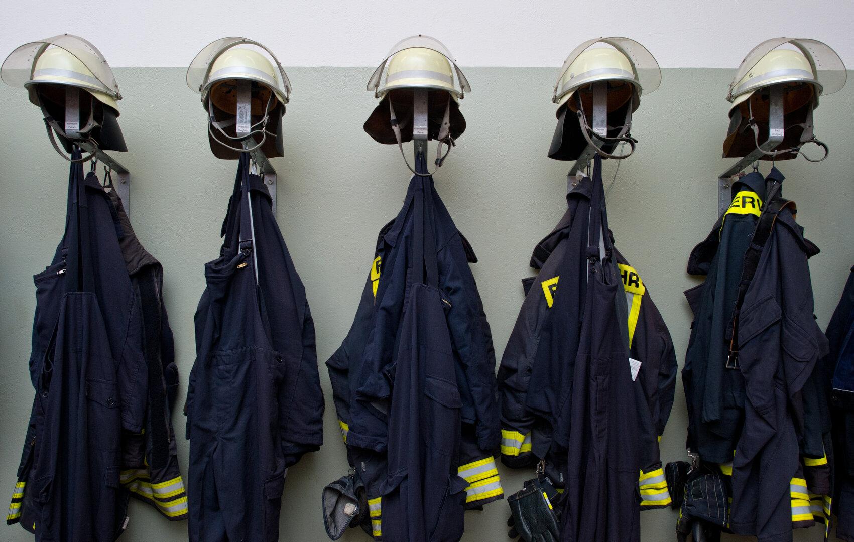 Mehrere Feuerwehrhelme und Uniformen hängen an Wandhaken in einer Reihe.