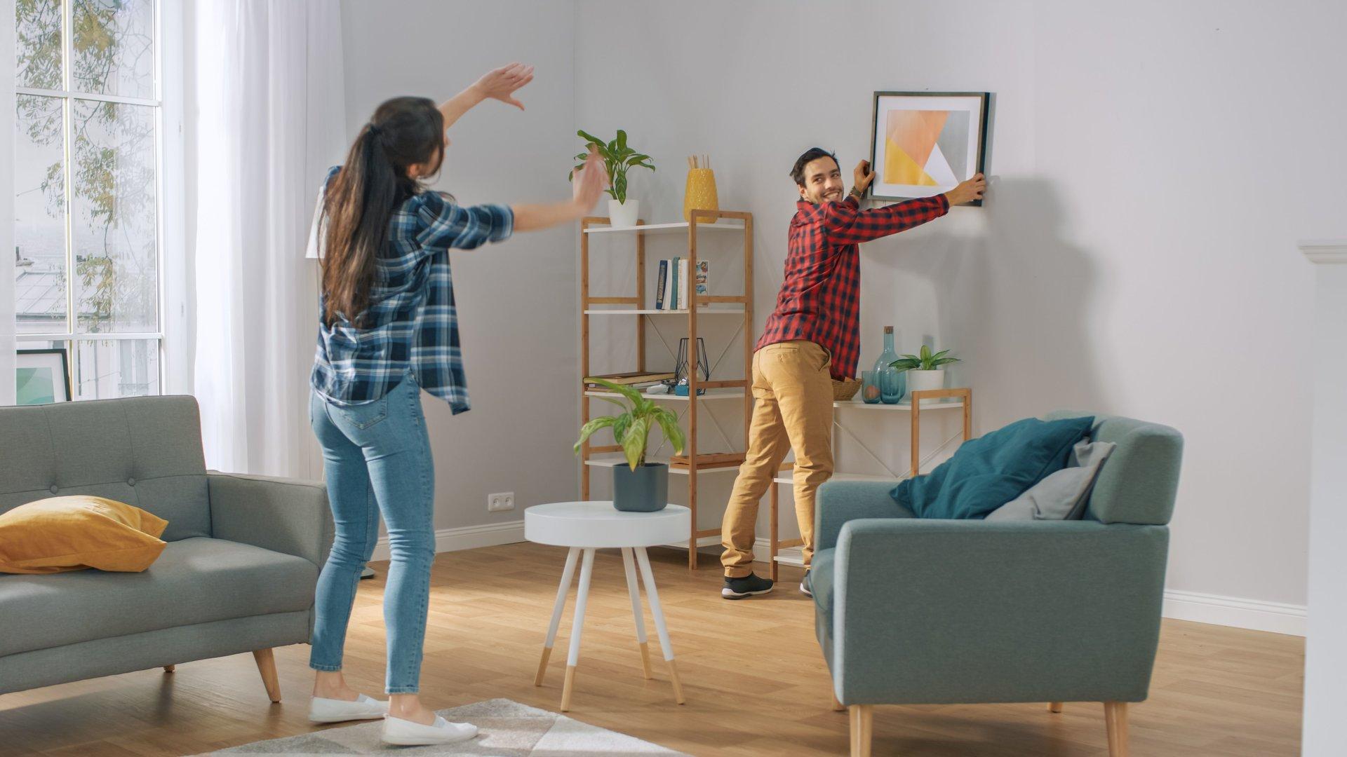 Happy Couple Hanging Picture on the Wall, Boyfriend Moves It, Girlfriend Tells Him when the Frame is Hanging Straight. Funny Moment in Young Couple's Life. Modern Stylish Apartment.