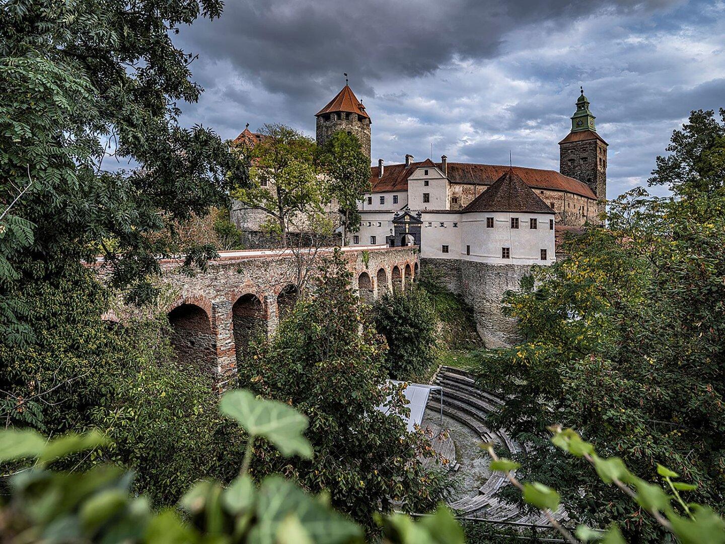 Burganlage mit Türmen, Brücke und Mauern über dichtem Grün unter düsterem Himmel.
