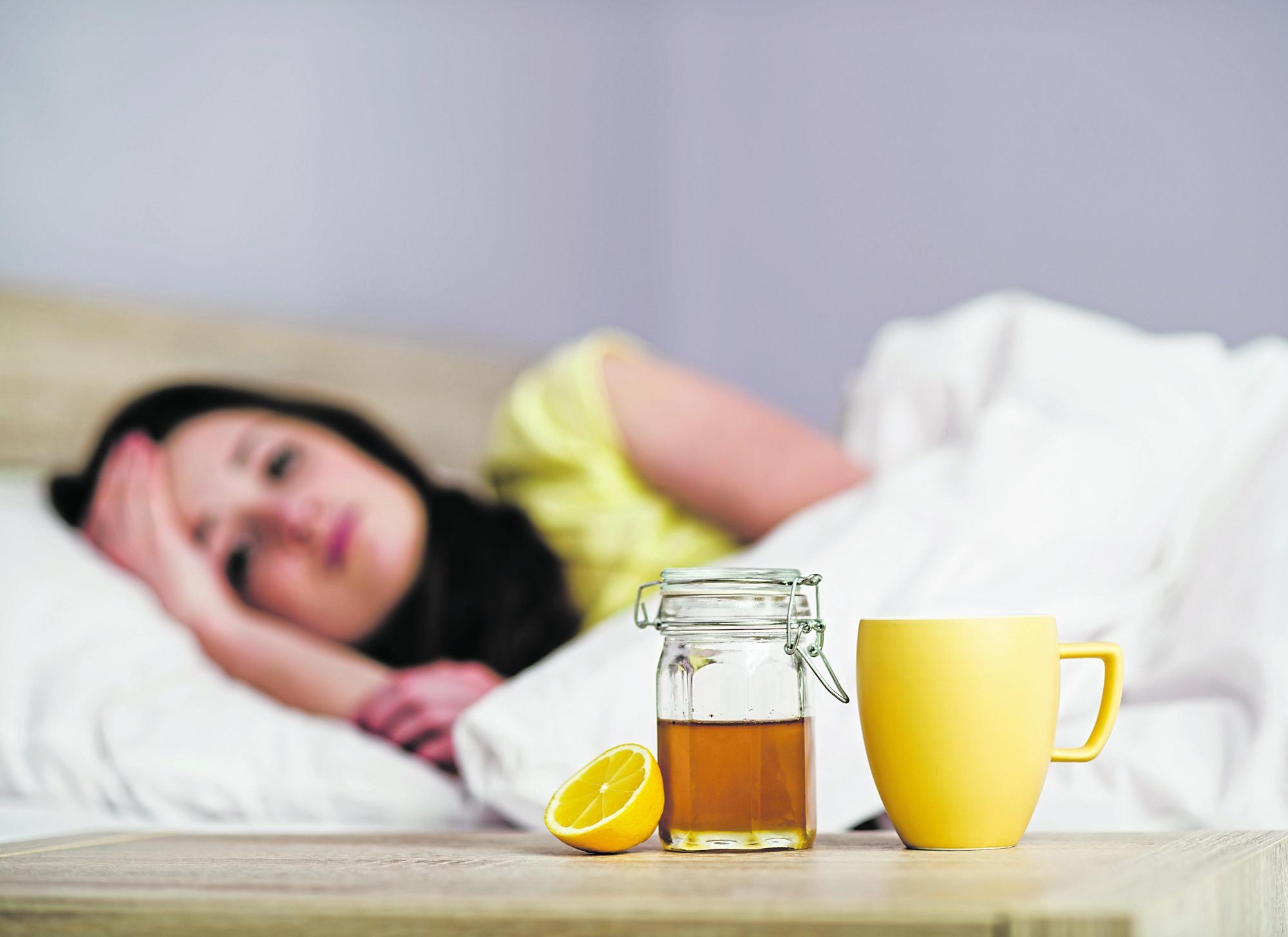 Frau mit Halsschmerzen liegt im Bett und hat die Hausmittel Honig, Zitronen und Tee bereitstehen.