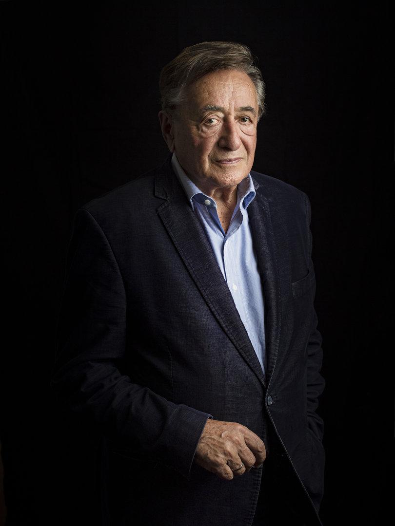 Ein älterer Mann in einem blauen Sakko vor einem schwarzen Hintergrund.