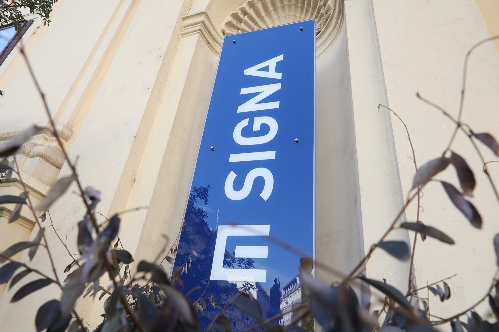 Ein blaues Schild mit der Aufschrift „SIGNA“ vor einem Gebäude.
