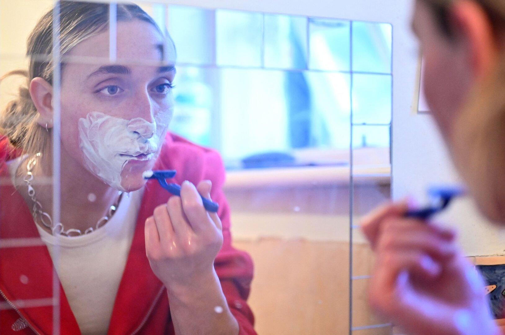 Eine Person mit Rasierschaum im Gesicht rasiert sich vor einem Spiegel.