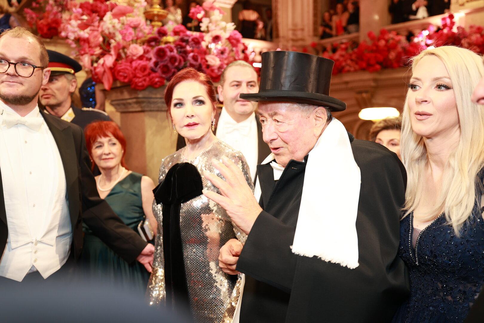 Prominente bei einer Veranstaltung, darunter ein Mann mit Zylinder und eine Frau im Paillettenkleid.