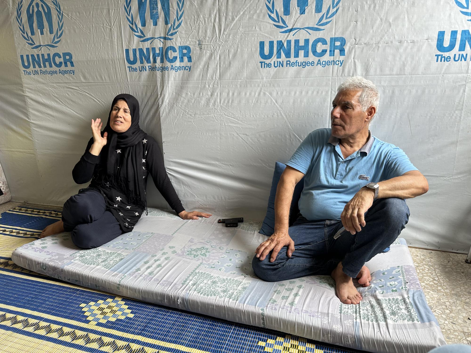 A woman in black clothes wearing a hijab sits on the floor, next to her a man in jeans and a polo shirt with no shoes on. Behind them is a grey courtain, the letters UNHCR are printed on it.